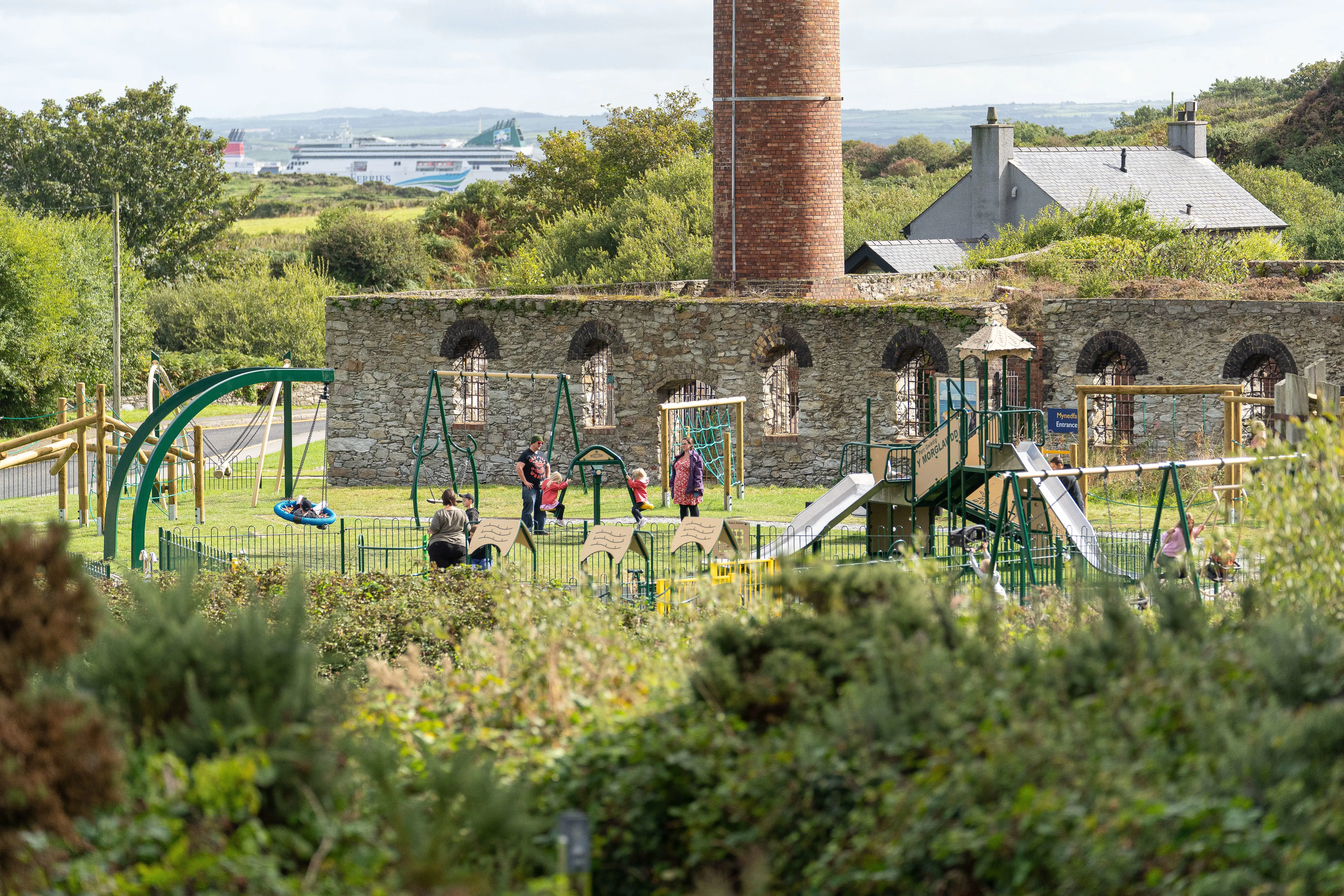 Parc chwarae newydd i blant gyda phlant ar swing ac oriel awyr agored yn y pellter