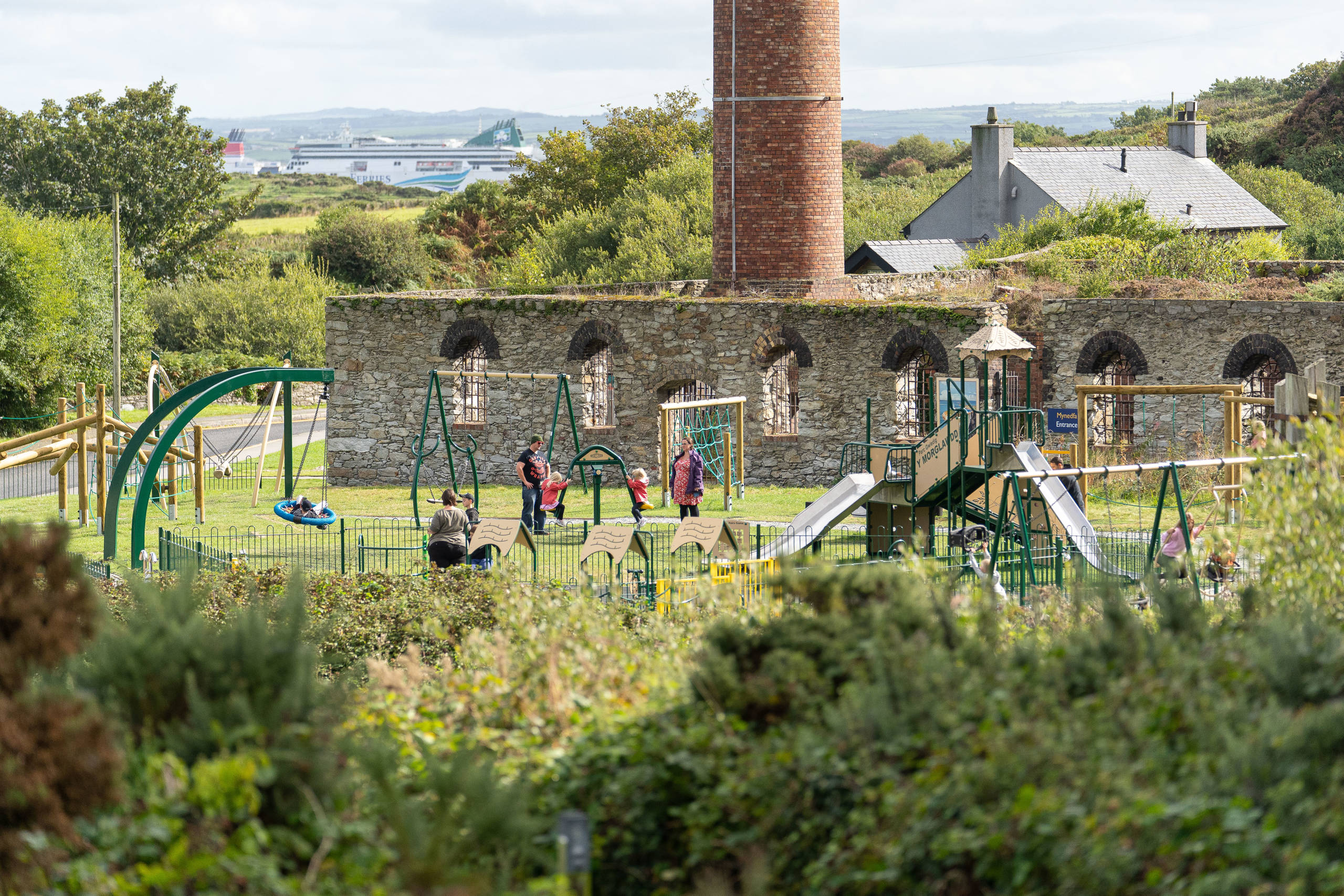 Parc chwarae newydd i blant gyda phlant ar swing ac oriel awyr agored yn y pellter