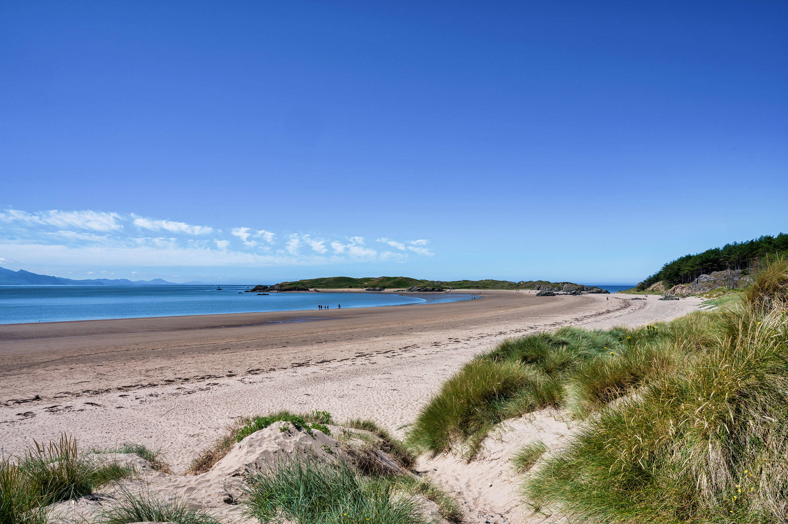 Bae Llanddwyn, yn dangos awyr las, traeth tywodlyd a thwyni tywod moresg gydag ynys Llanddwyn yn y pellter