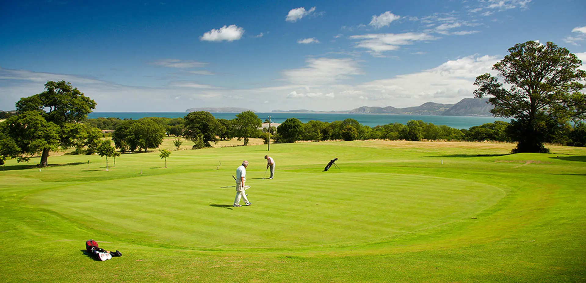 Dau berson yn chwarae golff ar y grîn olaf yn ceisio pytio gyda bag golff yn gorwedd ar y llawr