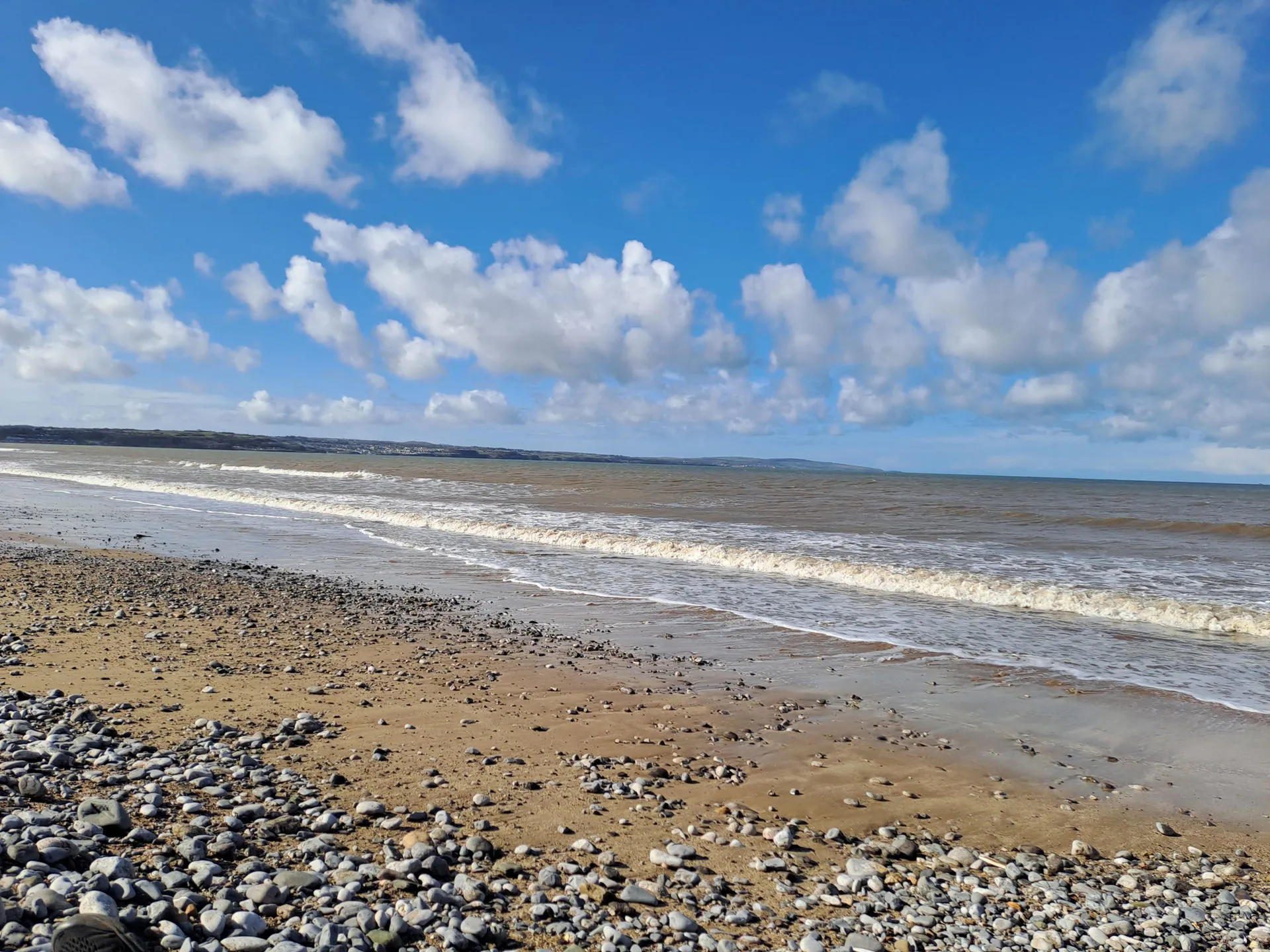 Golygfa yn edrych allan i'r môr gyda thonnau tonnog a cherrig mân o dan draed