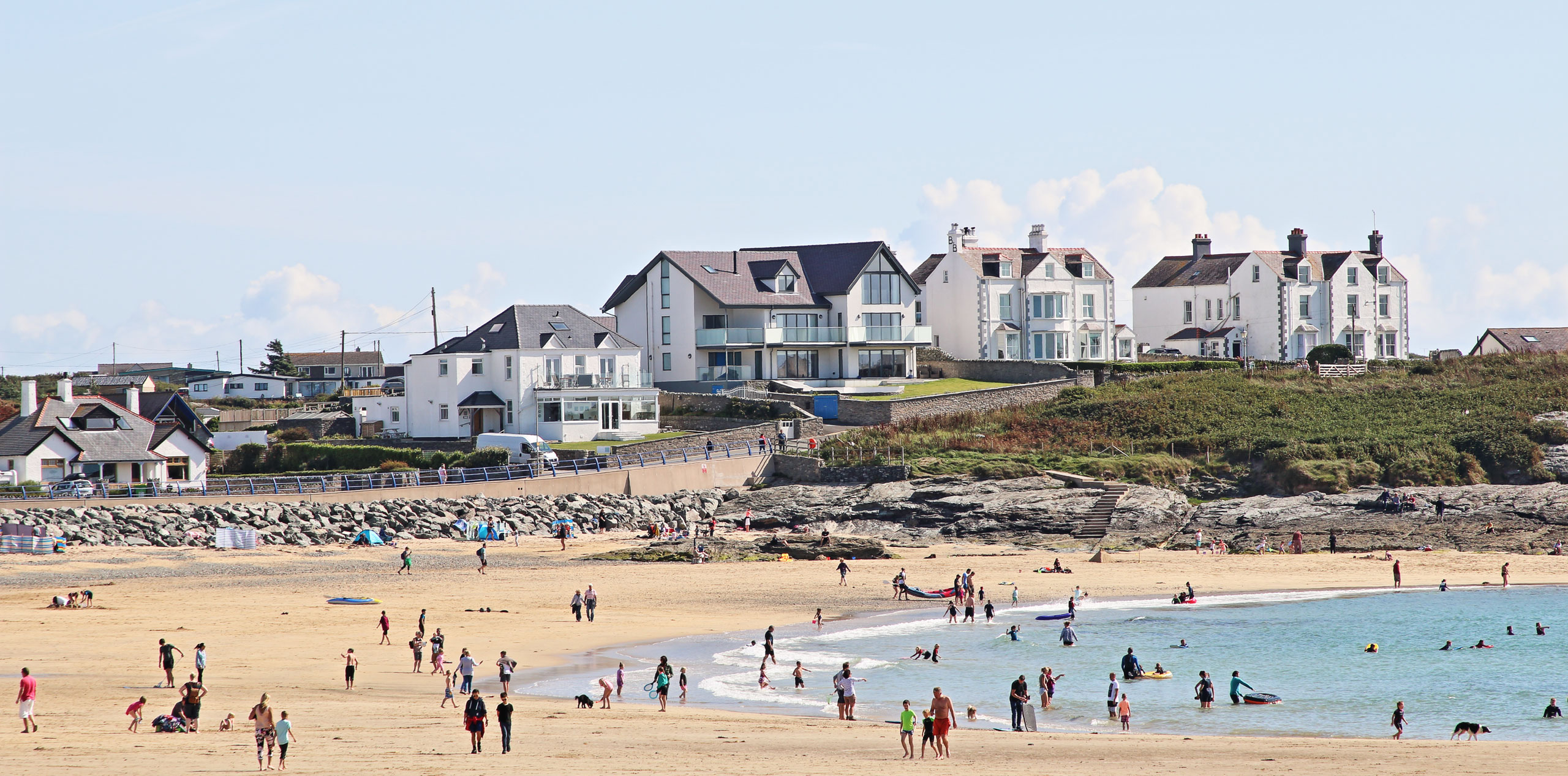 Tai yn y cefndir a diwrnod braf ar y traeth gyda pobl yn y môr ac ar y tywod
